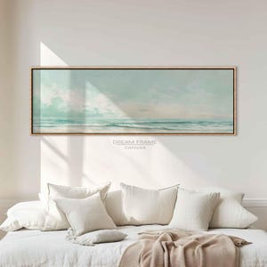 Puede incluir: Una impresión en lienzo rectangular de un paisaje marino en tonos azul claro y blanco. La obra representa el océano, las olas y el cielo. El lienzo está enmarcado en un marco fino de color marrón claro. El texto "DREAM FRAME CANVAS" está impreso en la parte inferior.
