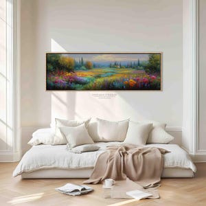 Puede incluir: Una pintura de paisaje enmarcada representa un campo vibrante de flores silvestres en tonos rojos, amarillos y morados. La obra de arte, con el texto "DREAM FRAME CANVAS", se exhibe sobre un sofá blanco con almohadas y una manta beige.