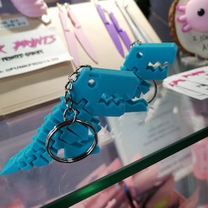 May include: A blue 3D printed dinosaur keychain. The dinosaur is made up of small, interlocking pieces. The keychain is attached to a silver ring.