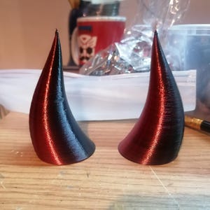 Può includere: Due corna di diavolo stampate in 3D, una rossa e nera, l'altra nera e rossa.