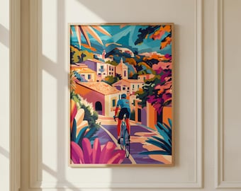 May include: A framed art print featuring a vibrant, stylized illustration of a cyclist riding a bicycle down a sunlit street in a Mediterranean-style village, surrounded by colorful buildings, lush pink flowers, and deep blue foliage under a bright sky.
