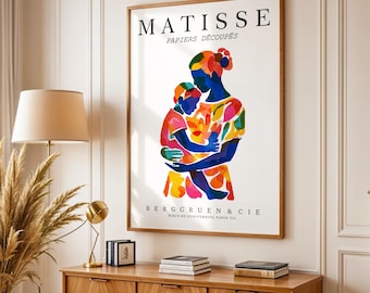 Może przedstawiać: Oprawiony wydruk artystyczny z kolorową ilustracją matki i dziecka obejmujących się. Grafika, zatytułowana "MATISSE PAPIERS DÉCOUPÉS", prezentuje żywe odcienie niebieskiego, czerwonego, żółtego i zielonego. Wydruk jest eksponowany na drewnianej szafce z lampą i elementami dekoracyjnymi.