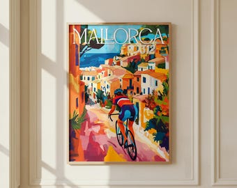 May include: A framed art print titled MALLORCA 312 featuring a colorful illustration of a cyclist riding down a sunlit street toward a coastal village with orange, yellow, and blue buildings, set against a deep blue sea and clear sky.