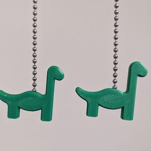 Puede incluir: Dos cadenas de tracción de color verde con forma de dinosaurio cuelgan de cadenas plateadas. Los dinosaurios están mirando en direcciones opuestas.