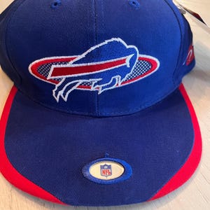 May include: A blue baseball cap with a red brim and a red button on top. The cap features the Buffalo Bills logo in white, red, and blue, and an NFL logo on the front.