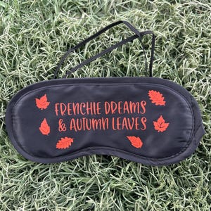 May include: A black sleep mask with the text "FRENCHIE DREAMS & AUTUMN LEAVES" in orange. The mask features orange leaf graphics and a black elastic strap. The mask is on green grass.