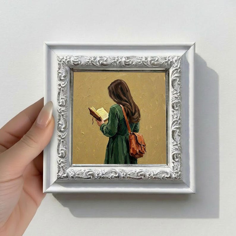 May include: A framed oil painting depicts a person in a green dress reading a book, with a brown satchel bag. The painting is set against a yellow background and framed in a white, ornate frame. The artwork is held by a hand.