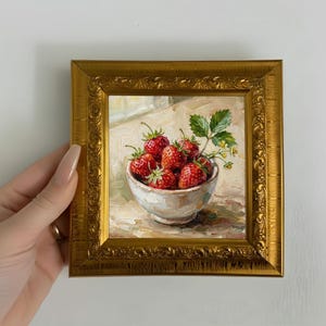 Puede incluir: Pintura al óleo enmarcada en un marco dorado y ornamentado que representa un cuenco blanco lleno de fresas rojas maduras. Se ven hojas verdes y pequeñas flores amarillas. La obra de arte es un bodegón.