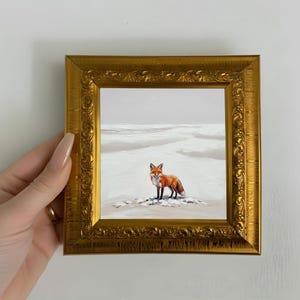 Puede incluir: Pintura enmarcada de un zorro en un paisaje nevado. El zorro tiene pelaje naranja y blanco y está sobre un parche de nieve. La pintura está en un marco dorado ornamentado.