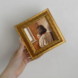 Op de afbeelding: Een kleine, vierkante, goudkleurige ingelijste schildering. Het kunstwerk toont een persoon met kort donker haar, gekleed in een witte trui, die een kopje vasthoudt bij een raam. De lijst heeft sierlijke details.