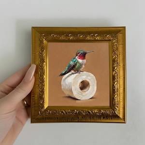 Può includere: Un piccolo dipinto in una cornice dorata ornata raffigurante un colibrì con piume verdi e rosse iridescenti appollaiato sopra un rotolo di carta igienica bianca su uno sfondo beige solido. L'opera cattura l'uccello di profilo.
