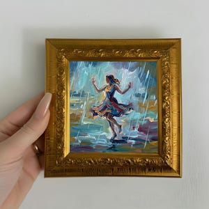 Op de afbeelding: Een ingelijst olieverfschilderij toont een vrouw die in de regen danst. Het kunstwerk heeft een gouden, sierlijke lijst. De vrouw draagt een kleurrijke jurk met tinten blauw, rood en geel. De achtergrond toont regen en een bewolkte lucht.