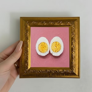 Könnte beinhalten: Gerahmtes Gemälde mit zwei Hälften eines hartgekochten Eies. Das Gemälde hat einen rosa Hintergrund und einen goldenen, verzierten Rahmen. Die Eigelb sind hellgelb und die Eiweiß sind weiß.