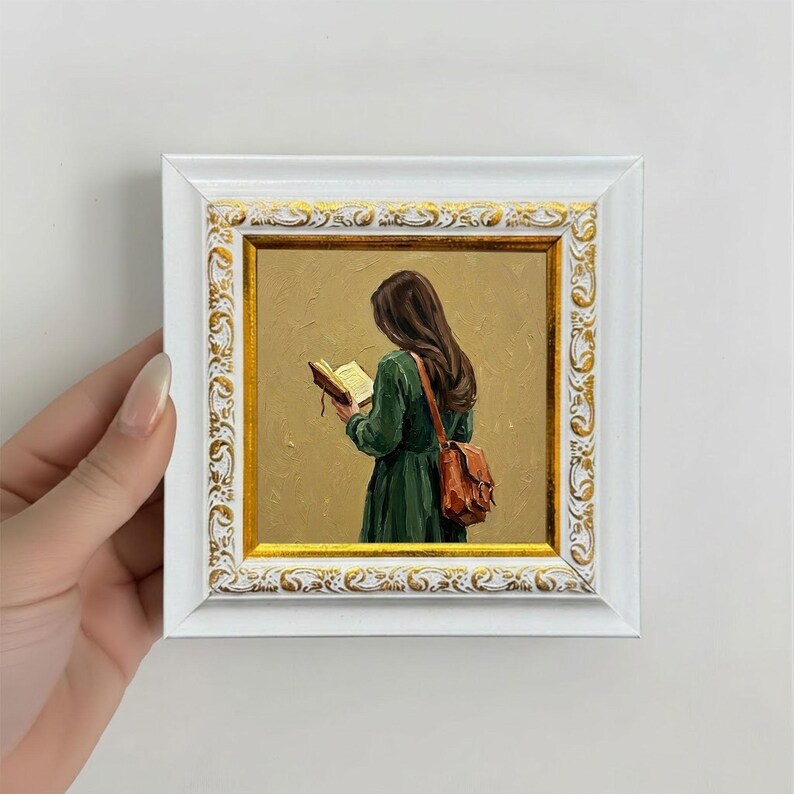 May include: A framed oil painting depicts a person in a green dress reading a book, with a brown leather bag. The painting is set against a gold background and framed in a white frame with gold accents. The artwork is held by a hand.