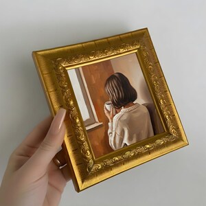 Op de afbeelding: Een schilderij in een gouden, sierlijke lijst. Het kunstwerk toont een persoon met kort bruin haar, gekleed in een witte trui, die een mok vasthoudt bij een raam. De achtergrond heeft warme tinten oranje en bruin.