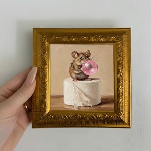 May include: A small square painting in an ornate gold frame depicting a brown mouse sitting on a white cylindrical object, blowing a large pink bubble. The mouse has detailed fur and whiskers, and the background is a solid, neutral beige color.