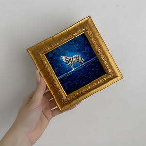 May include: A framed painting of a wolf howling at the moon. The artwork features a gold ornate frame and a dark blue background with a textured appearance. The wolf is depicted in shades of gray and white.