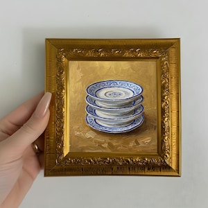 May include: A framed painting depicts a stack of four small, white plates with blue floral patterns. The artwork is set against a gold background and framed in a gold, ornate frame. The painting is held by a person.
