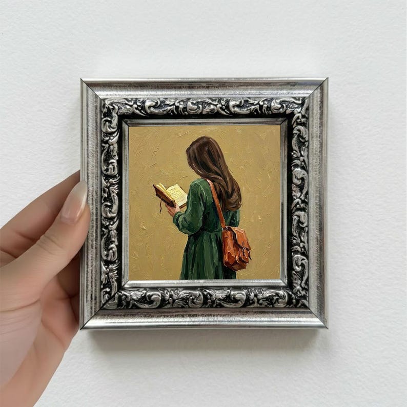 May include: A framed oil painting depicts a person in a green dress reading a book, with a brown leather satchel. The painting has a silver ornate frame. The background is a muted yellow.