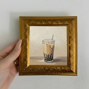 May include: A framed painting of a glass of bubble tea with a blue and white striped straw. The drink has a light brown color with dark tapioca pearls at the bottom. The frame is gold with ornate detailing.