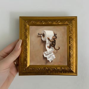Könnte beinhalten: Ein quadratisches Gemälde in einem verzierten Goldrahmen, das einen kleinen braunen Affen zeigt, der an einer Rolle weißem Toilettenpapier klettert, die an einem Wandhalter hängt. Das Kunstwerk hat einen neutralen beigen Hintergrund.