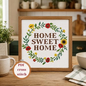May include: A framed cross-stitch featuring the words "HOME SWEET HOME" in brown, surrounded by a floral wreath of green leaves, red poppies, and yellow flowers. The frame is a light brown color. A small circular graphic reads "PDF cross stitch".