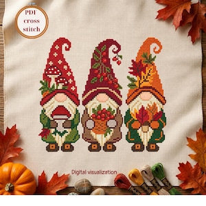 May include: A cross stitch pattern on fabric featuring three gnomes with autumn themes. The left gnome wears a red hat with mushrooms, the centre a dark red hat with berries, and the right an orange hat with maple leaves. Text reads PDF cross stitch.