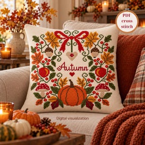 May include: A square throw pillow featuring a cross-stitch pattern with the word Autumn in red script, a large orange pumpkin, red mushrooms, acorns, and colourful autumn leaves arranged in a wreath design with a red bow at the top.