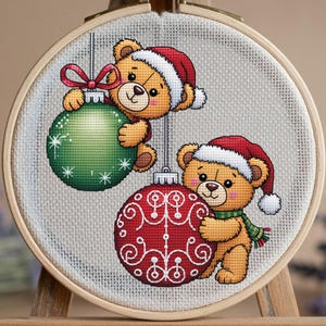 Può includere: Ricamo a punto croce di due orsetti con cappelli di Babbo Natale che tengono delle decorazioni natalizie. Uno ha un ornamento verde con fiocchi di neve bianchi, e l'altro un ornamento rosso con un disegno a spirale bianco. Il ricamo è in un telaio di legno.