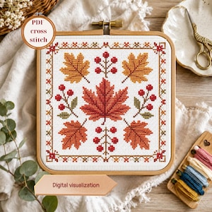 May include: A square wooden embroidery hoop containing a cross stitch pattern on white fabric. The design features a central red maple leaf surrounded by four smaller orange and red leaves and red berry sprigs, all framed by a decorative border.
