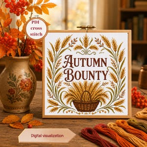 May include: A square wooden embroidery hoop containing a cross stitch pattern with the text Autumn Bounty in brown letters. The design features wheat stalks and a basket filled with wheat in shades of yellow, brown, and green on white fabric.