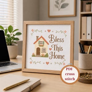 May include: A framed cross-stitch piece with the words "Bless This Home" and a house design. The house features a red roof, a beige facade, and a brown door. The frame is made of light-colored wood. A PDF cross stitch logo is in the bottom right corner.