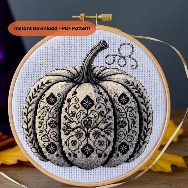May include: A cross-stitch pattern of a pumpkin in a wooden embroidery hoop. The pumpkin is detailed with black floral designs and a curved stem. The text "Instant Download &bull; PDF Pattern" is in an orange banner at the top.