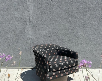 May include: A black barrel chair with a patterned design of small squares in red, silver, and white. The chair has a rounded back and sits on a brown, circular base. The chair is outdoors against a gray wall.