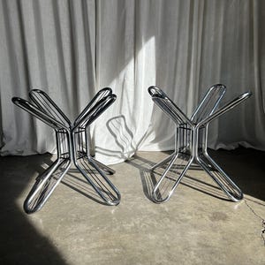 May include: Two chrome metal table bases. Each base has a starburst design with curved legs and a series of parallel bars forming the support structure. The bases are on a concrete floor with a white curtain backdrop.