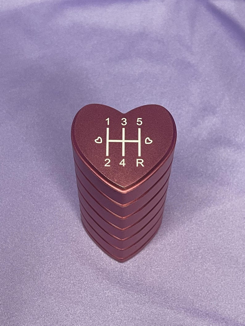 K&ouml;nnte beinhalten: Ein herzf&ouml;rmiger, metallisch roter Schaltknauf. Die Oberseite des Knaufs zeigt das Gangmuster: 1, 3, 5 und R, mit den Zahlen 2 und 4 darunter. Der Knauf hat ein geschichtetes Design und liegt auf einer violetten Oberfl&auml;che.