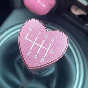 Könnte beinhalten: Rosa herzförmiger Schalthebelknauf mit weißen Zahlen und Gängen. Das Schaltschema ist mit R, 1, 2, 3, 4, 5 und 6 beschriftet. Ein passender rosa wolkenförmiger Knopf ist im Hintergrund zu sehen.