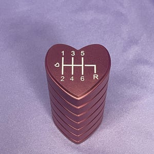 K&ouml;nnte beinhalten: Ein herzf&ouml;rmiger, metallisch roter Schaltknauf. Das Schaltungsmuster ist in Wei&szlig; dargestellt: 1, 3, 5, 2, 4, 6 und R. Der Knauf hat ein gestapeltes Design mit einer wiederholten Herzform.