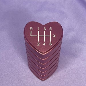 K&ouml;nnte beinhalten: Ein herzf&ouml;rmiger, metallisch roter Schaltknauf. Die Oberseite des Knaufs zeigt das Schaltschema in Wei&szlig;, mit den Buchstaben "R" und den Zahlen "1, 3, 5" oben und "2, 4, 6" unten. Ein kleines Herzsymbol befindet sich neben der Zahl 6.