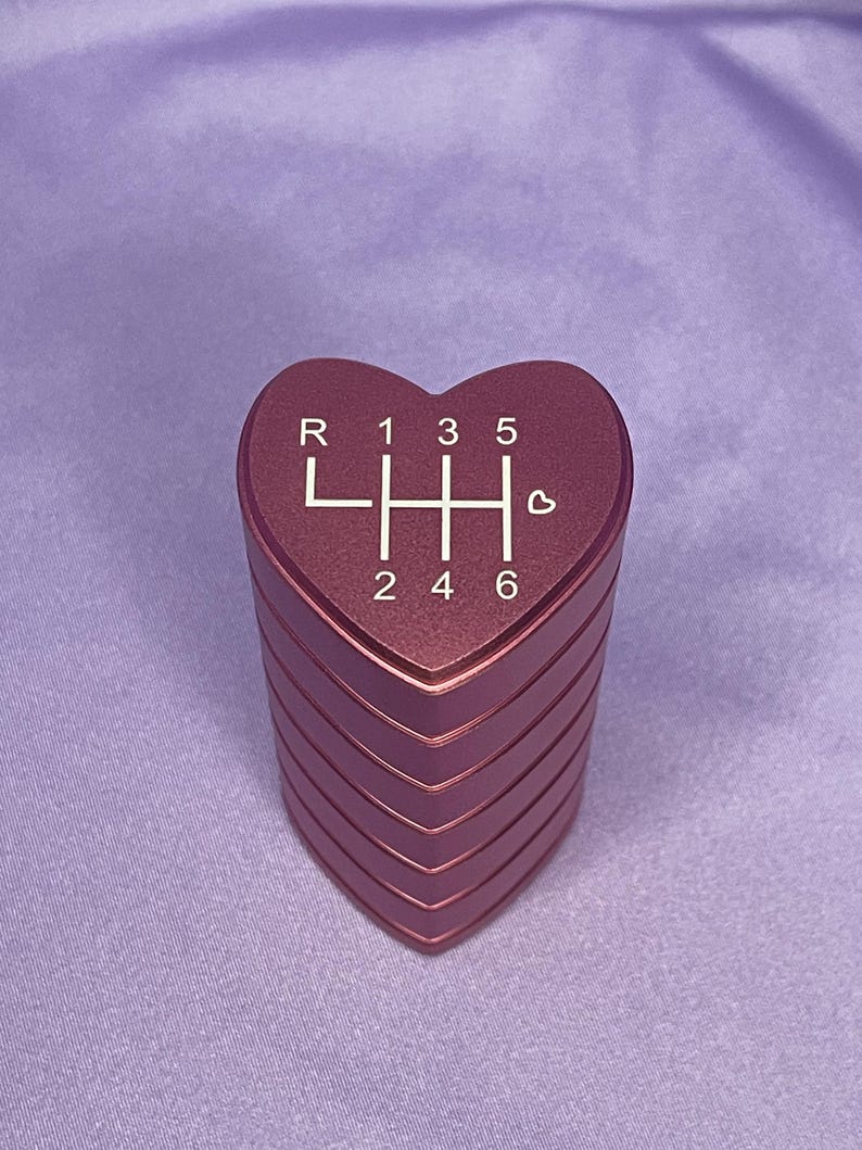 K&ouml;nnte beinhalten: Ein herzf&ouml;rmiger, metallisch roter Schaltknauf. Die Oberseite des Knaufs zeigt das Schaltschema in Wei&szlig;, mit den Buchstaben "R" und den Zahlen "1, 3, 5" oben und "2, 4, 6" unten. Ein kleines Herzsymbol befindet sich neben der Zahl 6.