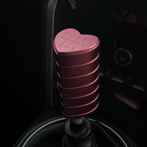 K&ouml;nnte beinhalten: Ein rosa, herzf&ouml;rmiger Schaltknauf mit geripptem Design. Die Oberseite des Knaufs zeigt das Schaltschema in Wei&szlig;. Der Knauf ist im Innenraum eines Autos installiert, mit einer schwarzen Lederschaltmanschette.