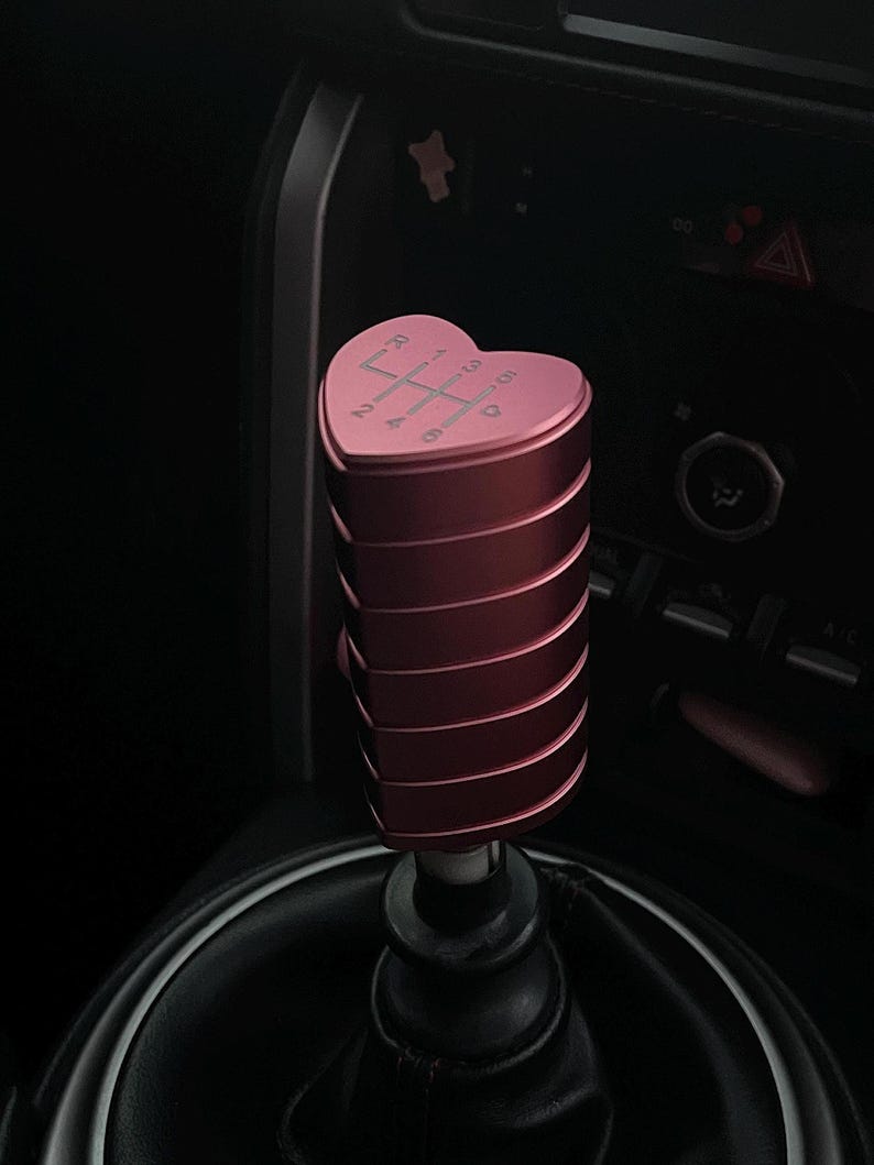 K&ouml;nnte beinhalten: Ein rosa, herzf&ouml;rmiger Schaltknauf mit geripptem Design. Die Oberseite des Knaufs zeigt das Schaltschema in Wei&szlig;. Der Knauf ist im Innenraum eines Autos installiert, mit einer schwarzen Lederschaltmanschette.