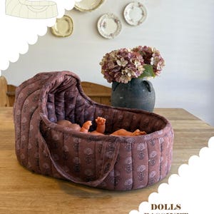 Op de afbeelding: Een gewatteerde poppenwieg van oudroze stof met een herhalende donkere bloemenprint. De wieg heeft een kap en geïntegreerde handvatten, geschikt voor poppen van 38cm en kleiner. Tekst: PERFECT FOR DOLLS 38CM AND SMALLER, DOLLS BASSINET PDF sewing pattern, avalen horn patterns.