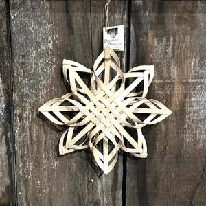 May include: A white woven star ornament, hanging from a string. The star is made of thin strips of wood, woven together in a complex pattern. The star is hanging against a wooden background. The tag on the star reads "Designer Baskets by Donna".
