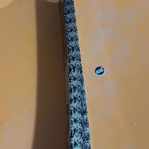 May include: A cricket bat with a white grip and a black and gold patterned blade. The bat has a sticker with the letters "KBS" on it. The bat is lying on an orange surface.