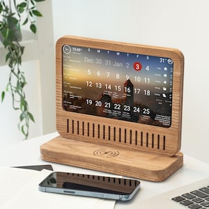 Può includere: Un calendario e orologio digitale in legno che mostra la data, l'ora e il meteo. Il calendario ha una base in legno con un pad di ricarica wireless. Uno smartphone è posizionato davanti al calendario.