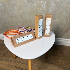 Puede incluir: Regleta de madera con múltiples enchufes y puertos USB. La regleta es rectangular e inclinada, con un acabado de madera clara. También hay dos regletas verticales de madera, cada una con múltiples enchufes y puertos USB. Se ven cables naranjas y morados.
