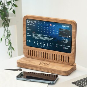 Puede incluir: Una pantalla digital de madera que muestra la fecha, la hora, el clima y datos financieros. La pantalla se encuentra sobre una base de madera con una plataforma de carga inalámbrica. Un teléfono inteligente descansa frente a la pantalla.