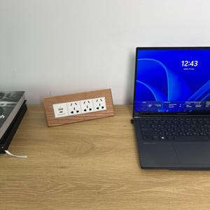 Puede incluir: Un enchufe de madera con cuatro enchufes y un puerto USB. El enchufe está colocado sobre un escritorio de madera junto a un portátil con una pantalla azul que muestra la hora 12:43. Una pila de libros está a la izquierda.
