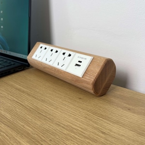 Könnte beinhalten: Eine Holz-Steckdosenleiste mit vier Steckdosen und zwei USB-Anschlüssen. Die Steckdosenleiste besteht aus hellem Holz und hat eine weiße Frontplatte mit dem Text "DC 5V MAX 10W". Sie liegt auf einem Holzschreibtisch.